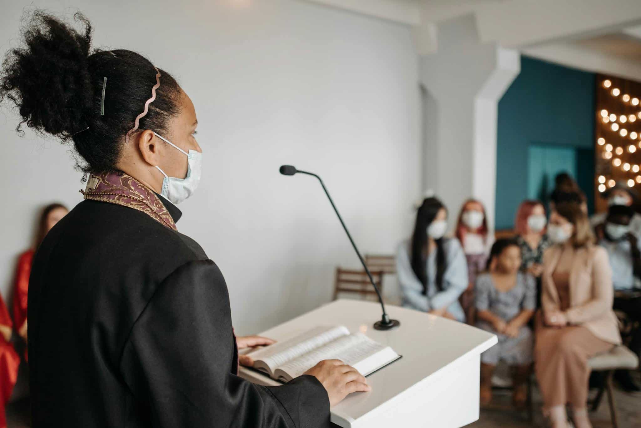 Female pastor delivering a sermon to a masked congregation indoors.